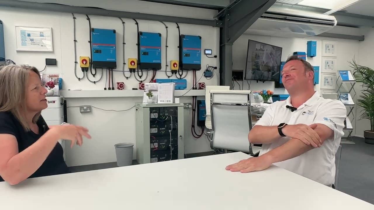 Twee mensen in gesprek aan een tafel voor een wand vol Victron omvormers/acculaders — videobeeld