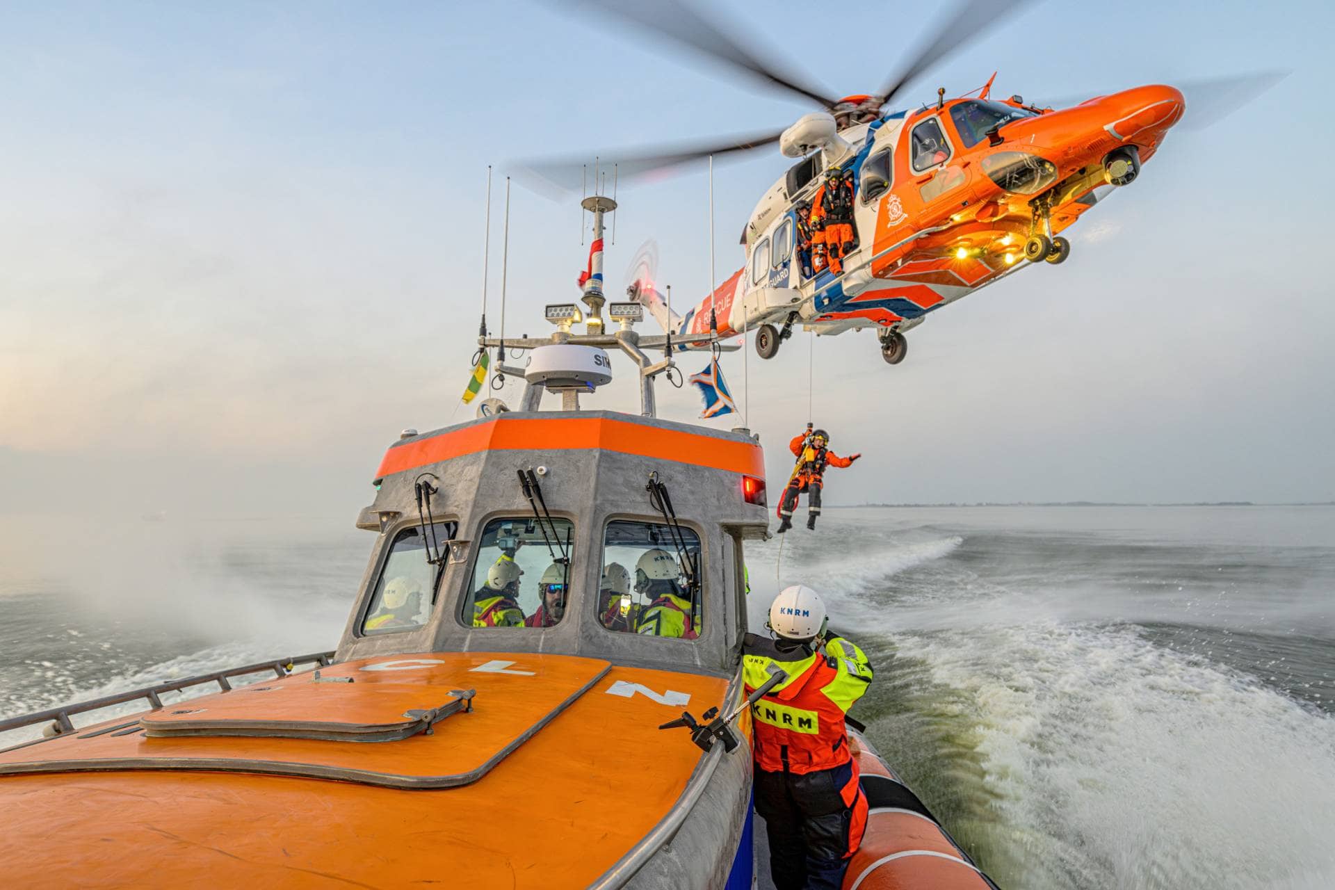 KNRM-reddingsboot op zee met een reddingshelikopter die een bemanningslid omhoog takelt